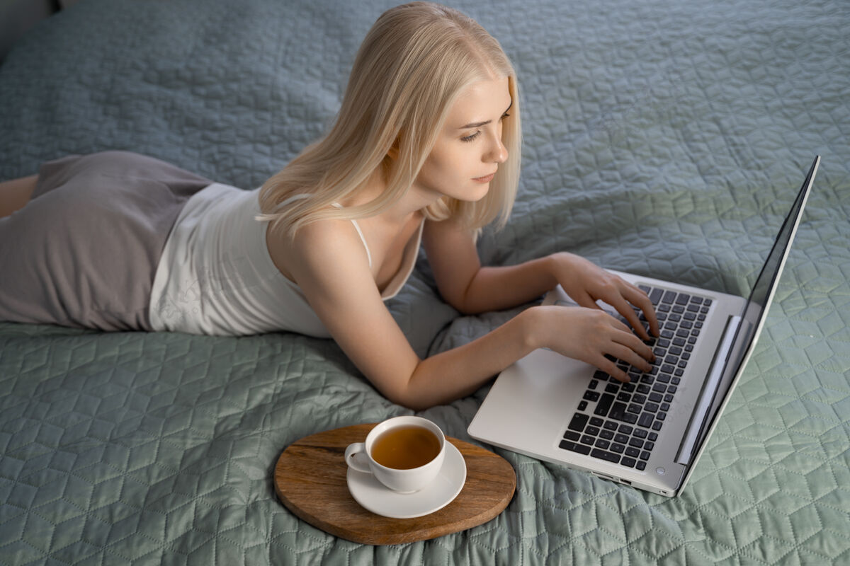 床一个随意的金发女郎在家里的床上使用笔记本电脑的画像女孩放松笔记本电脑