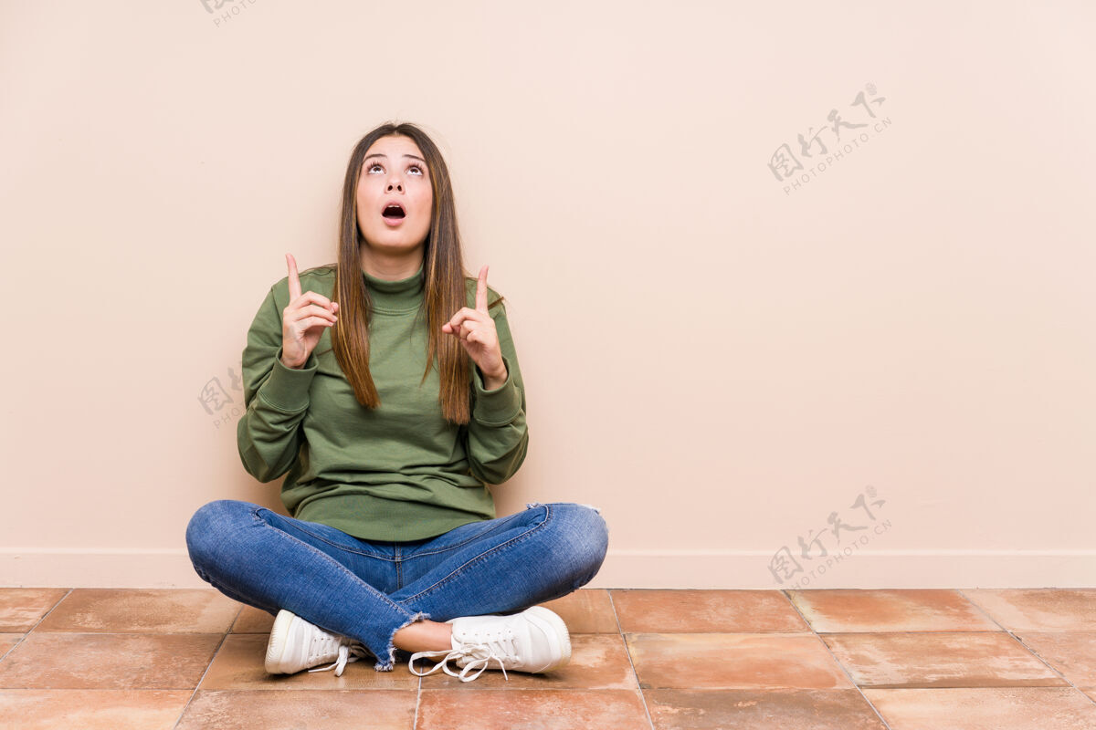 快乐年轻的白人妇女坐在地板上 张着嘴 孤立地指着上面产品自信哇