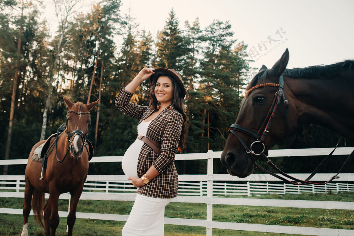 肚子一个大肚子的怀孕女孩 戴着帽子 挨着一个围场附近的马自然动物户外