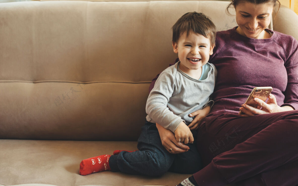 聊天一对白人母亲和儿子坐在沙发上微笑着使用手机的正面照片父母笑聪明