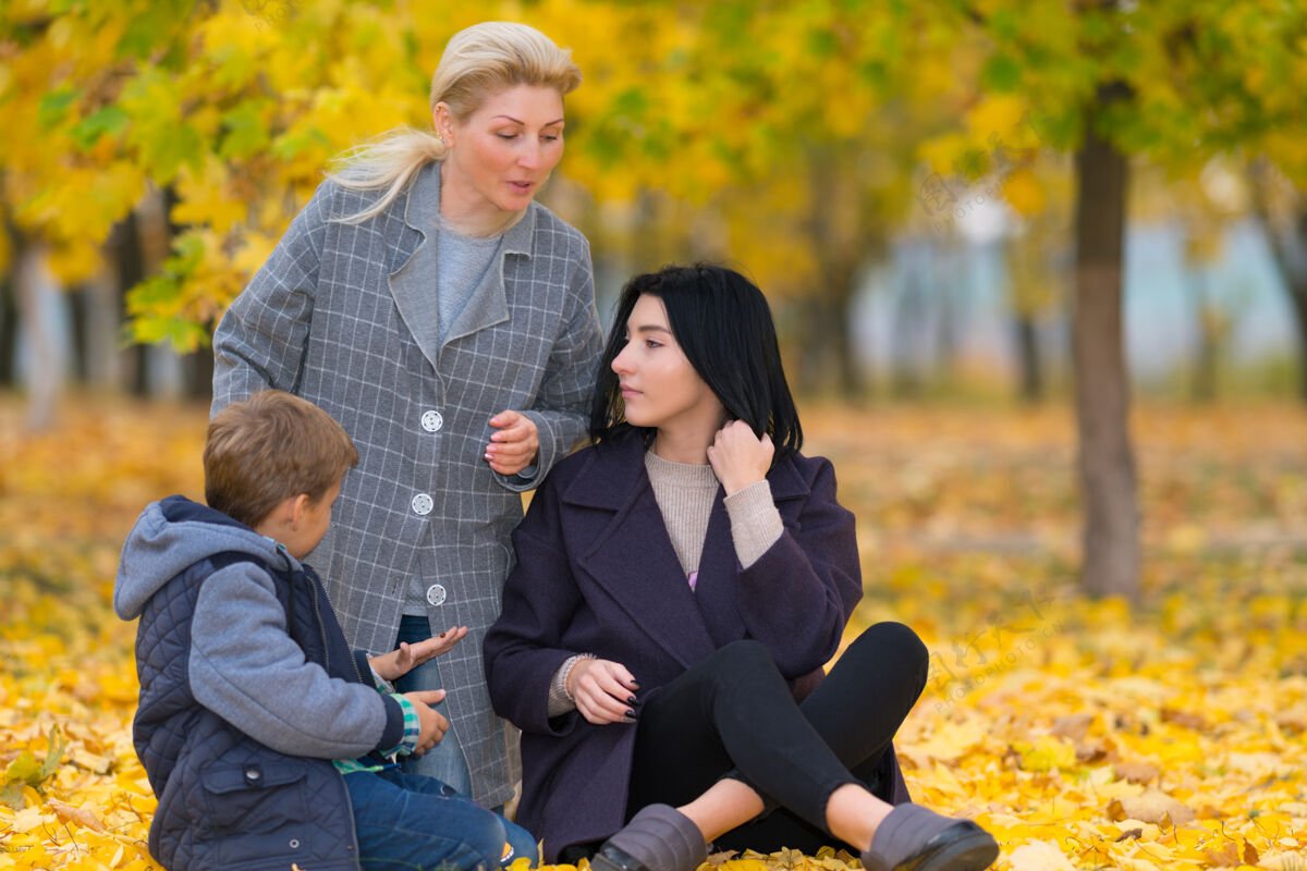 儿子秋天公园里的一个年轻家庭坐在地上聊天 在有树的林地里 五颜六色的黄叶中间未成年陪伴债券
