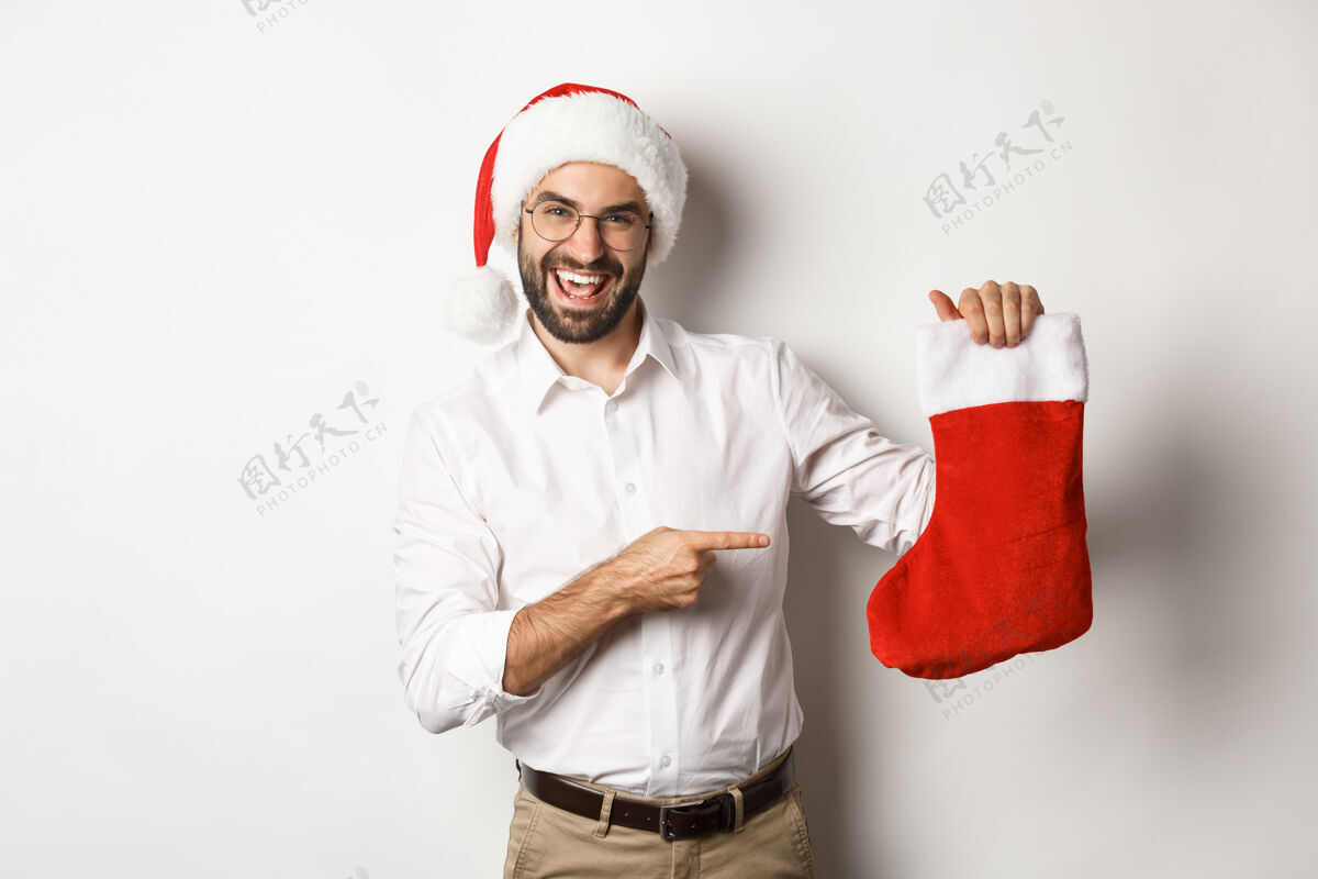 成人戴着圣诞帽庆祝寒假的快乐男人 指着圣诞袜微笑着 拿着礼物 站在白色的背景上圣诞老人商人微笑