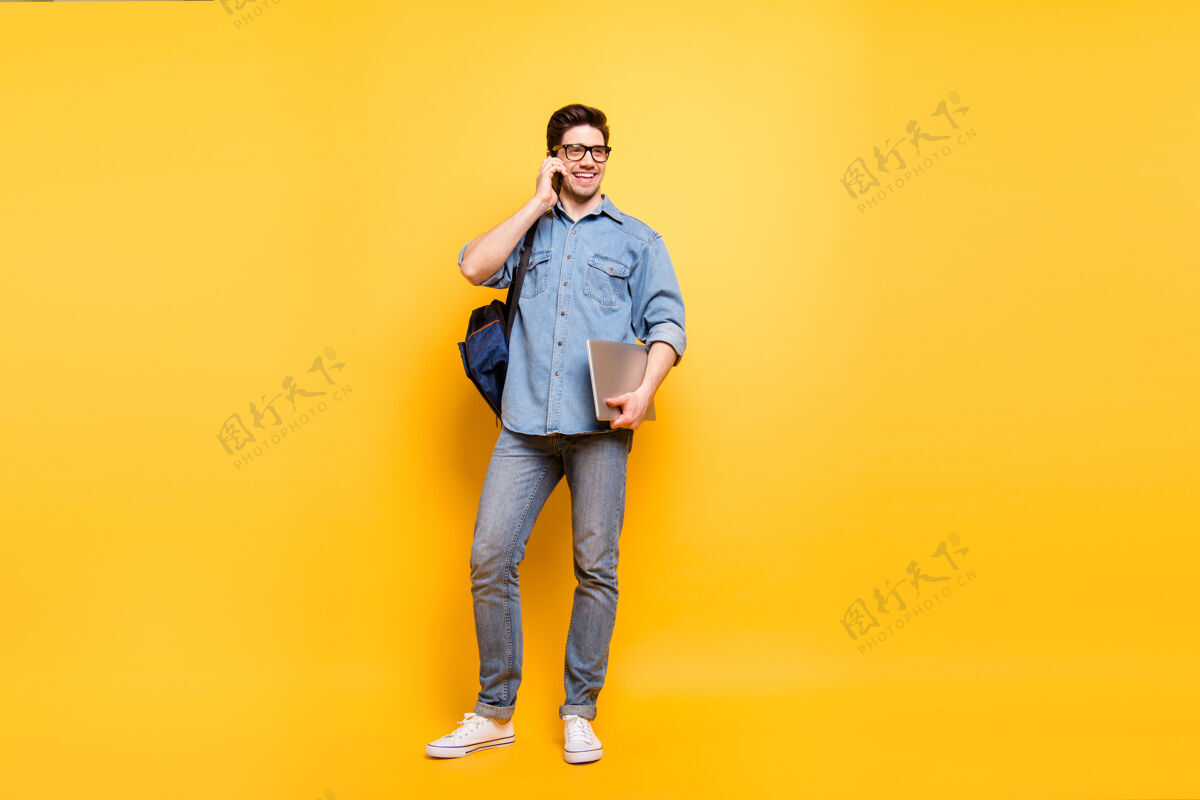 通话一张全身尺寸的照片 照片上是一个快乐的 积极的 满嘴笑容的男人 手里拿着笔记本电脑 穿着白色运动鞋打电话 隔着一堵充满活力的彩色墙学习交谈背包