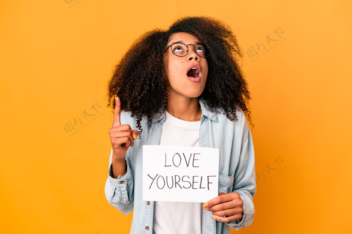 女性年轻的非洲裔美国卷发女子举着一个“爱你自己”的标语牌 张大嘴巴指向上方女人美国产品