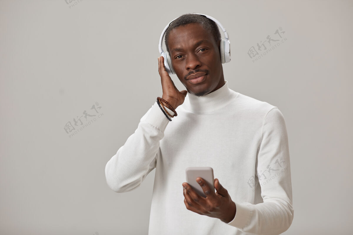 连接照片中的年轻人戴着无线耳机用手机听音乐隔离在灰色的墙上自信手持微笑