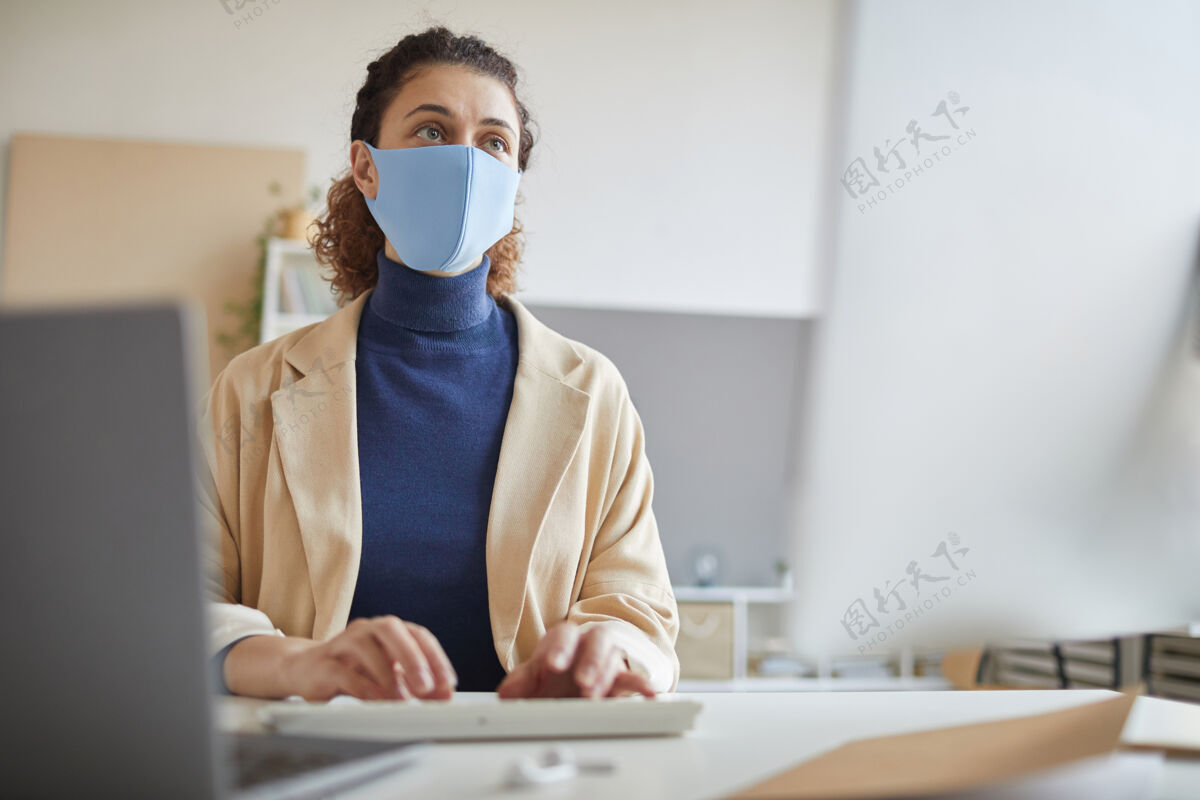 公司业务戴着防护面具的年轻女子看着电脑显示器 她在大流行期间上网工作办公室忙碌无线技术