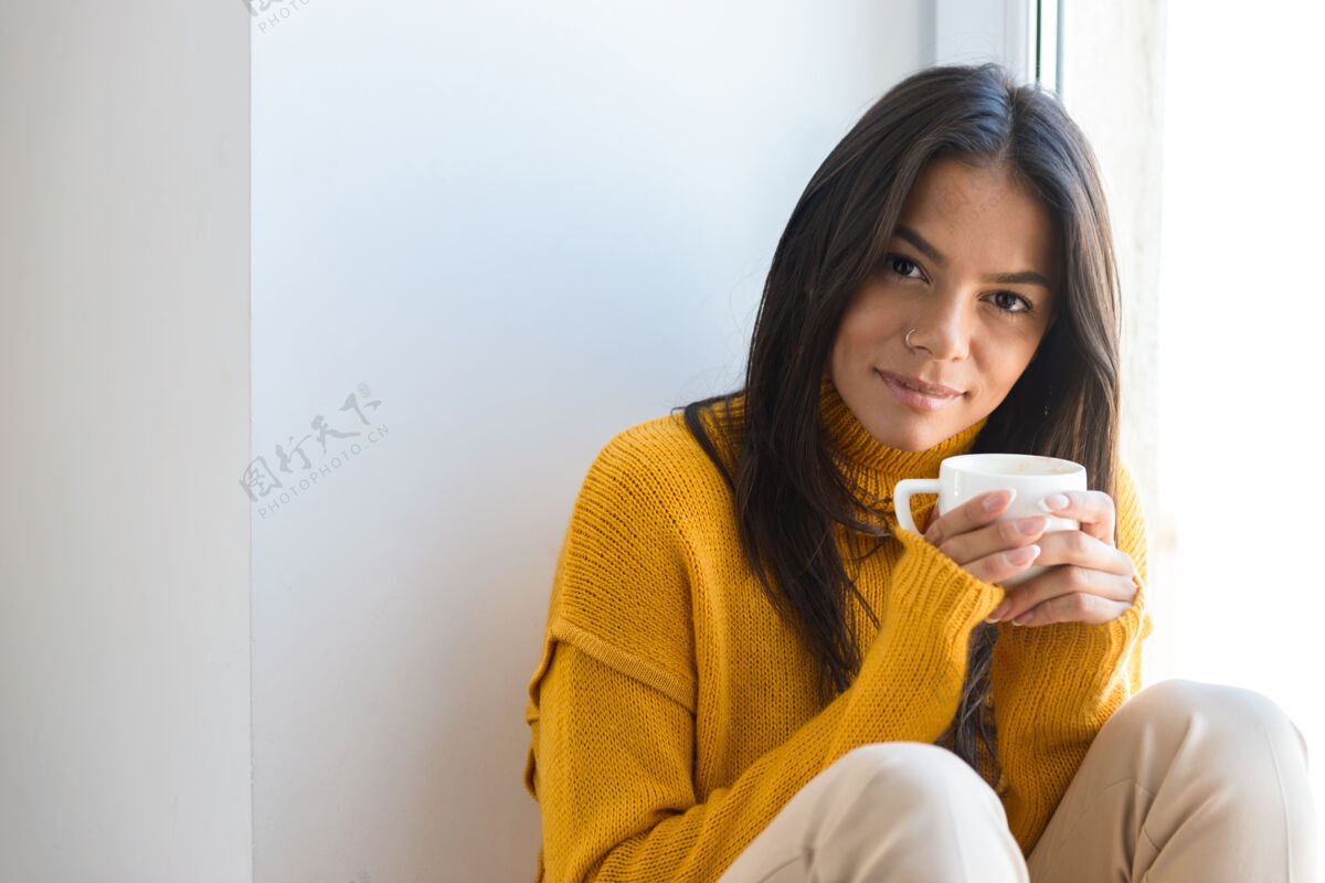 冬天一个穿着毛衣的可爱的年轻女子坐在室内的窗边 端着一杯茶的特写镜头季节茶微笑