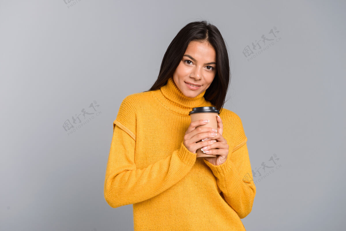 看起来一个微笑的年轻女子穿着毛衣 拿着外卖咖啡杯 隔着灰色的墙吸引力茶手臂