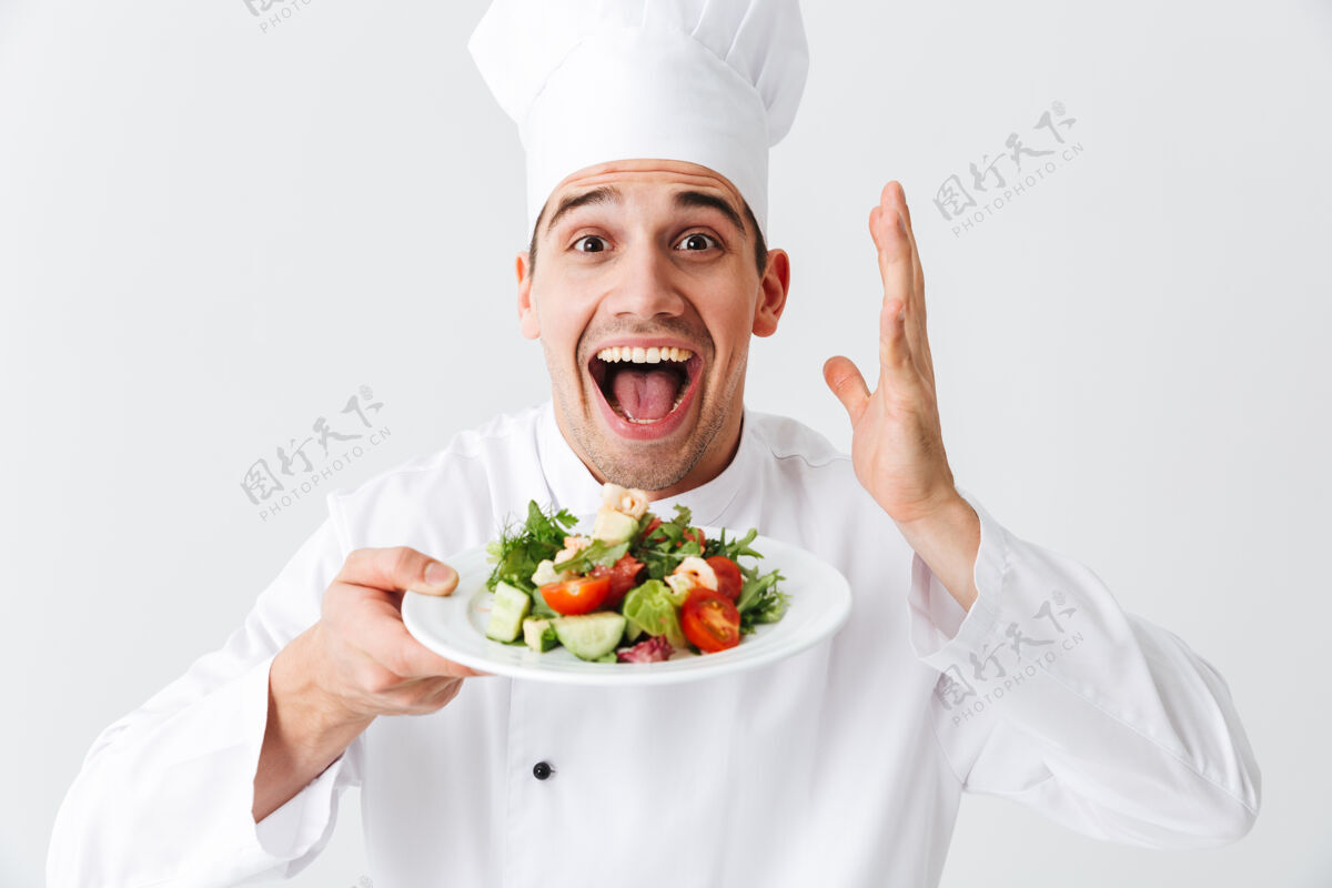 信心兴奋的男厨师穿着制服 把新鲜的蔬菜沙拉放在隔离在白色墙上的盘子上午餐服务高加索