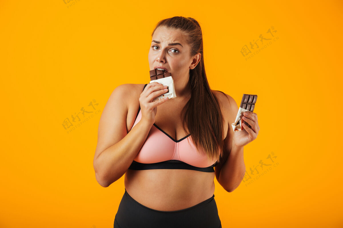 年轻人一个穿着运动服的快乐超重的年轻女子孤立地站在黄色的墙上 吃着巧克力女人女孩高兴