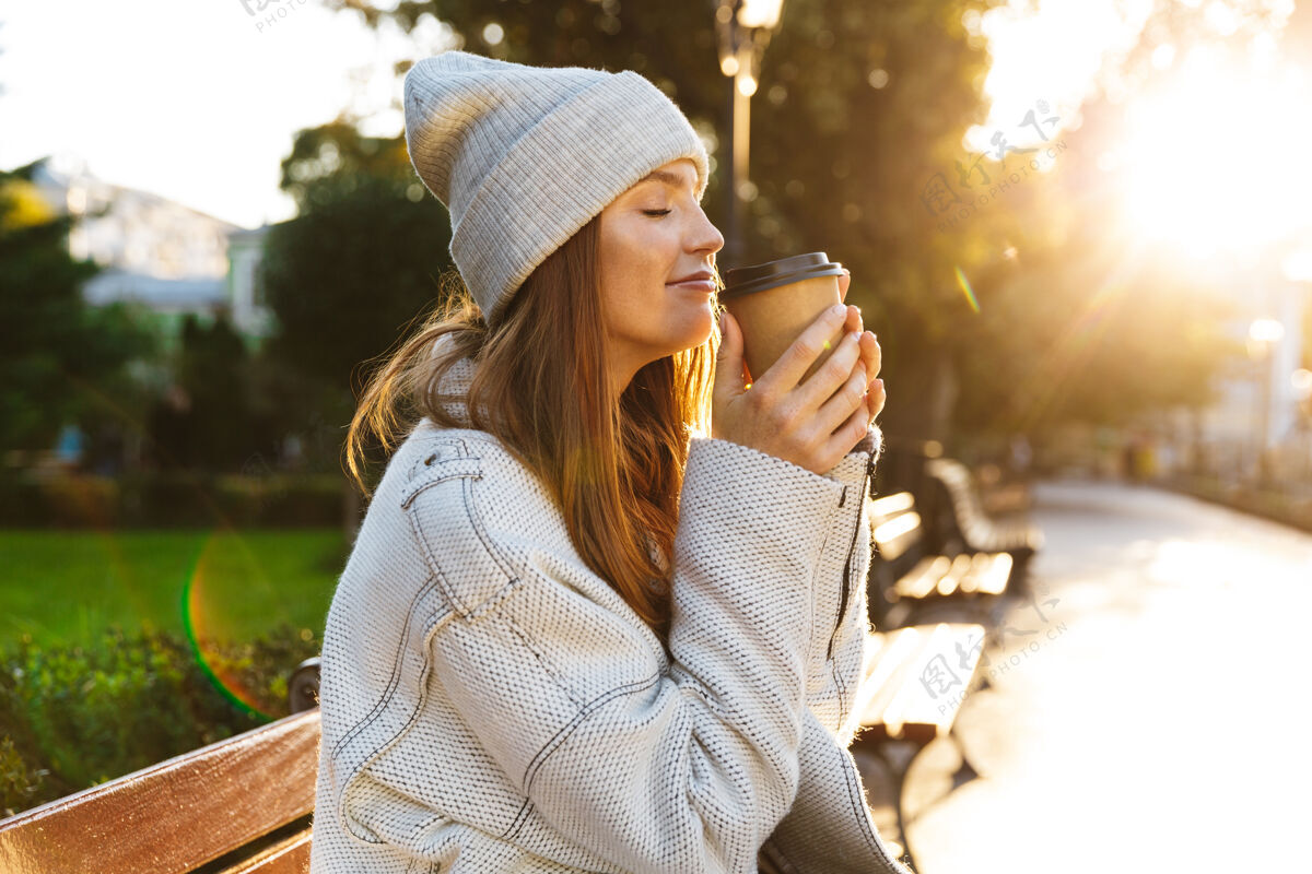 坐着美丽的年轻女子穿着秋大衣 戴着秋帽 坐在户外的长凳上 端着一杯热咖啡看时尚宁静