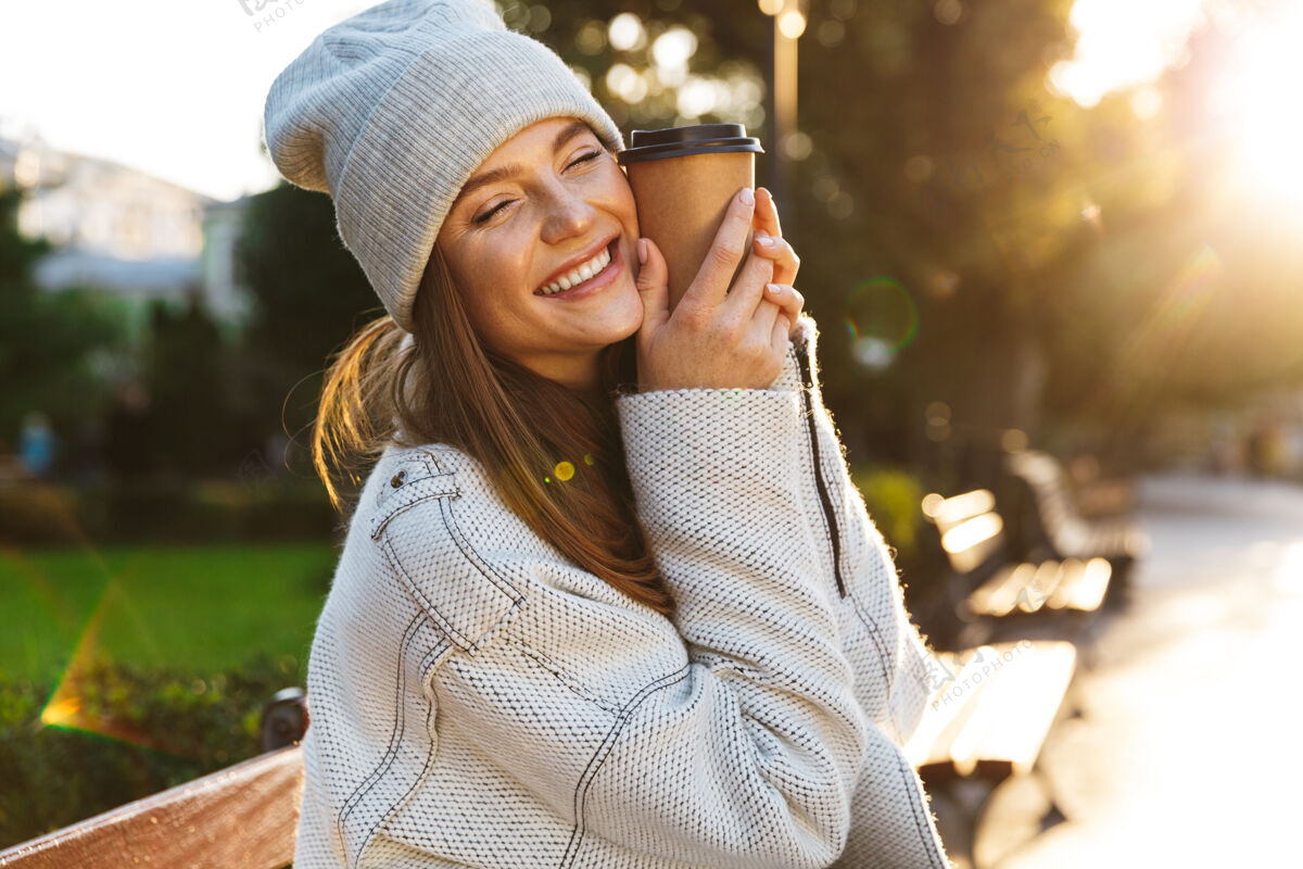 饮料美丽的年轻女子穿着秋大衣 戴着秋帽 坐在户外的长凳上 端着一杯热咖啡季节热人
