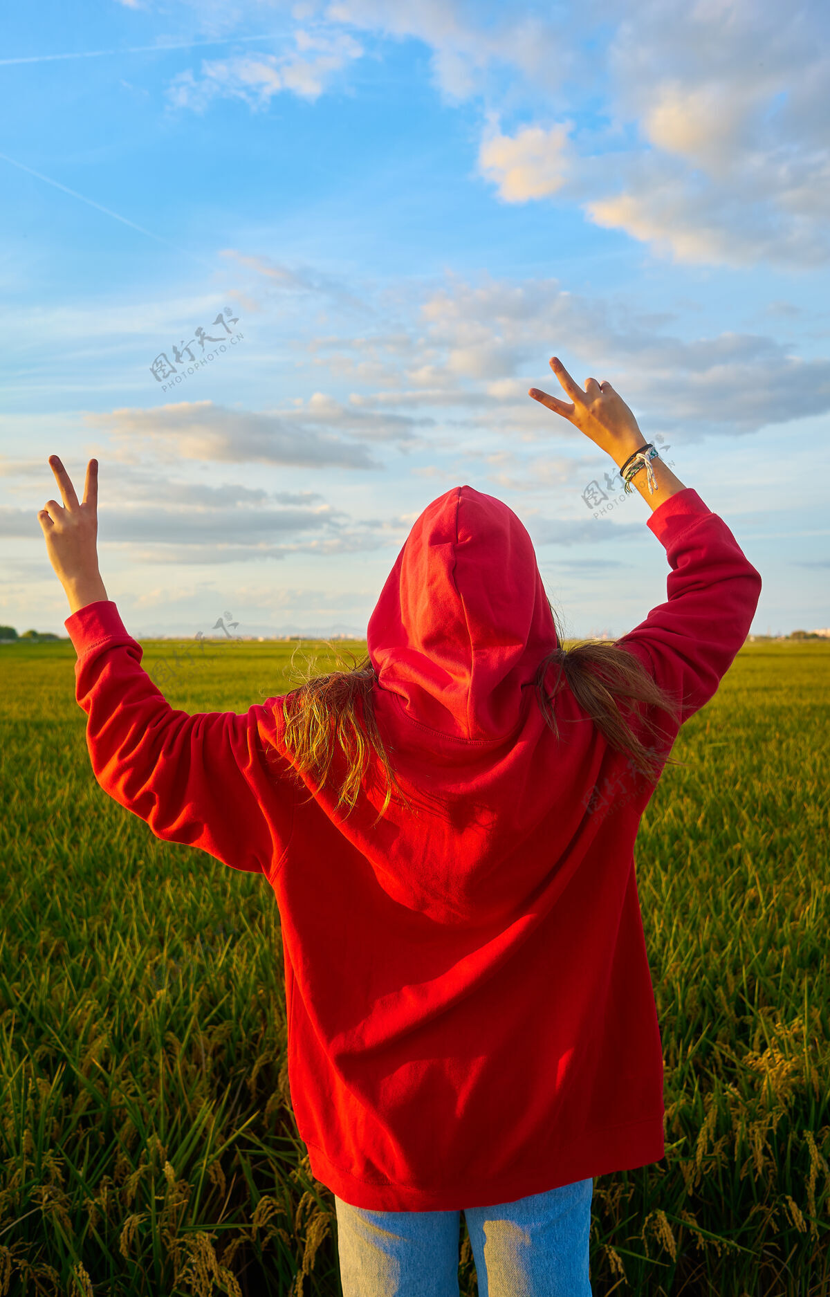 时尚日落时分 一个身穿红色夹克 头戴红帽的小女孩在稻田里用手做着胜利的手势时髦女孩姿态