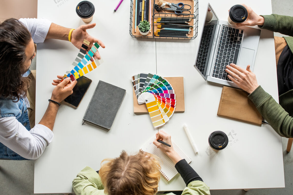 伙伴关系三个年轻的创意设计师坐在办公桌旁 从调色板上为新项目选择颜色 其中一个正在做笔记工程师办公室会议