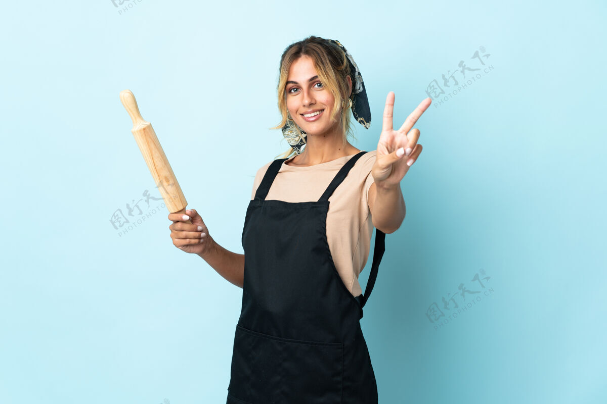 烹饪年轻的金发乌拉圭厨娘孤立在蓝色微笑和显示胜利的标志工作女人人