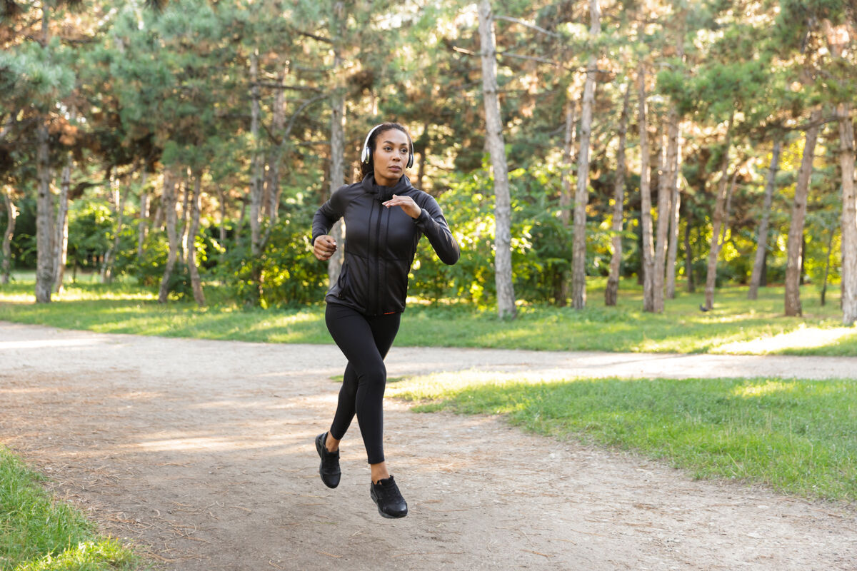 年轻一个20多岁的漂亮女人穿着黑色运动服 戴着耳机 一边在绿色公园里跑步跑步温暖伸展
