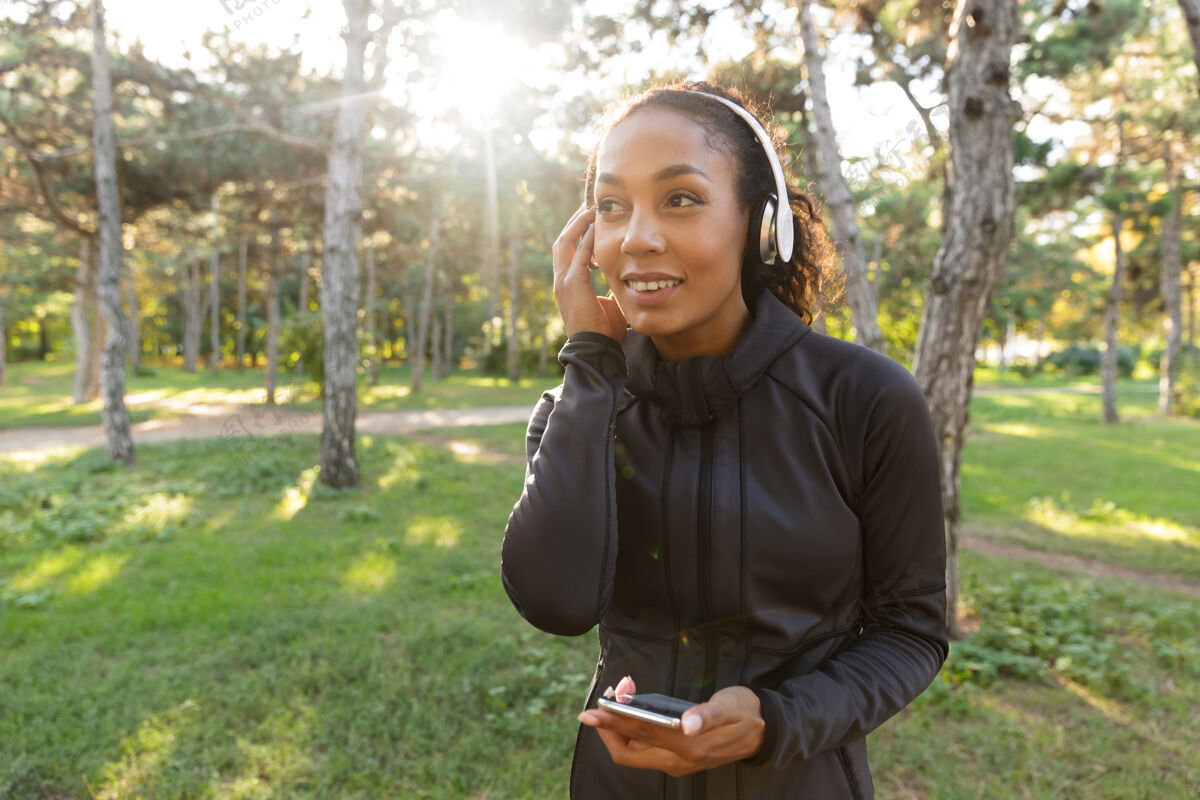 减肥20多岁的快乐女人穿着黑色运动服 戴着耳机 一边在绿色公园里散步一边用手机女性运动员公园