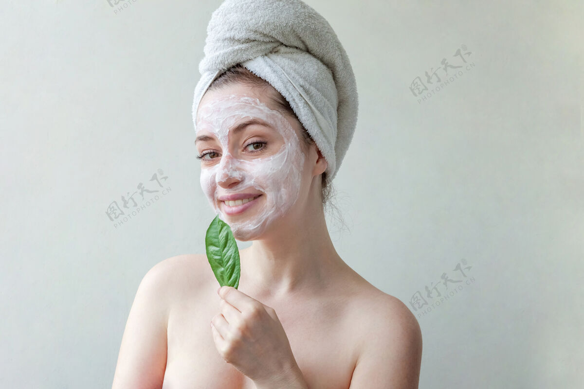 卫生头戴毛巾 脸上涂有白色滋养面膜或乳霜 手拿绿叶 白色背景的美女肖像治疗护理皮肤科