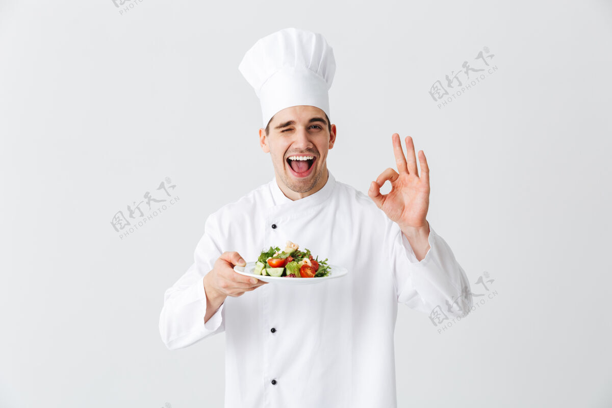 帽子兴奋的男厨师穿着制服 在白色墙壁上展示一盘新鲜的蔬菜沙拉 好吧烹饪举行食物