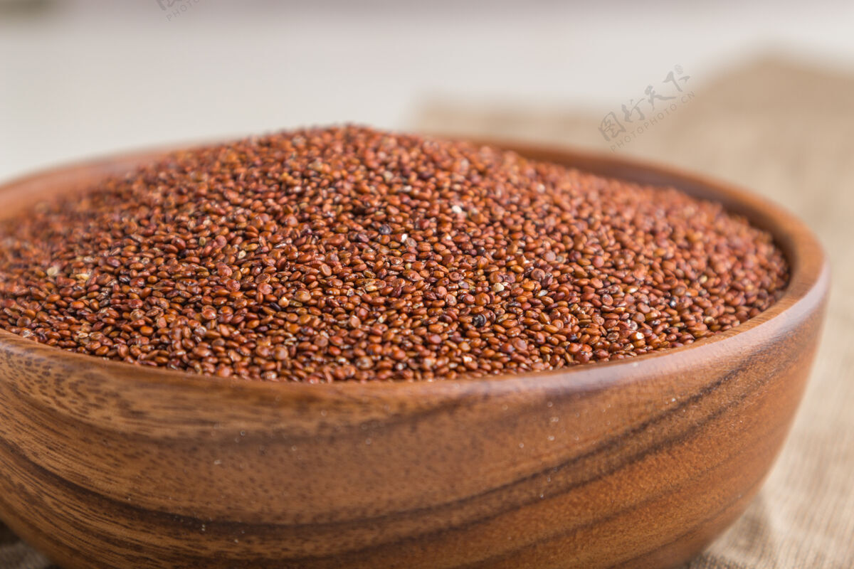 干白色木质表面和亚麻织物上有生的红色奎奴亚藜种子的木碗勺子碗美食