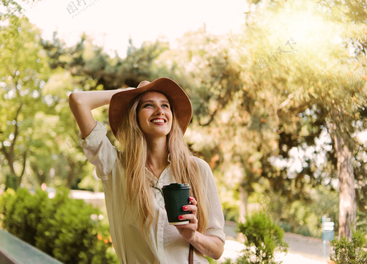 电话开朗的金发女人穿着休闲风格的衣服 戴着毡帽 边走边喝咖啡 在城市里散步微笑年轻人时尚
