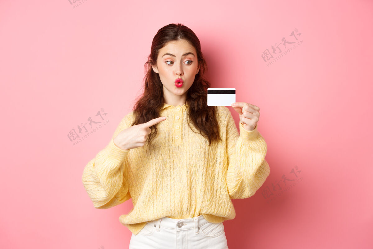 黑发迷人的女孩去购物 看起来很兴奋 指着塑料信用卡 站在粉红色的墙上情绪旁白请