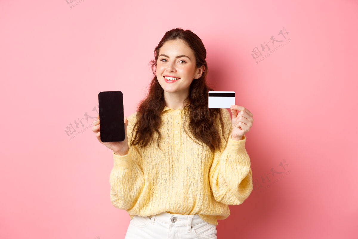 在线科技和网上购物美丽女士的肖像展示手机屏幕和塑料信用卡 微笑着高兴 推荐应用程序 站在粉红色的墙上电话女性信用卡