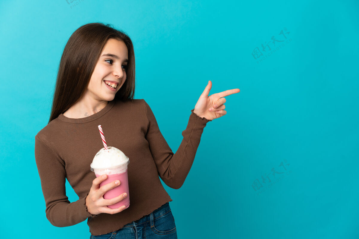 牛奶拿着草莓奶昔的小女孩被隔离在蓝色的墙上 手指指向一边 展示一个产品演示表情眼睛