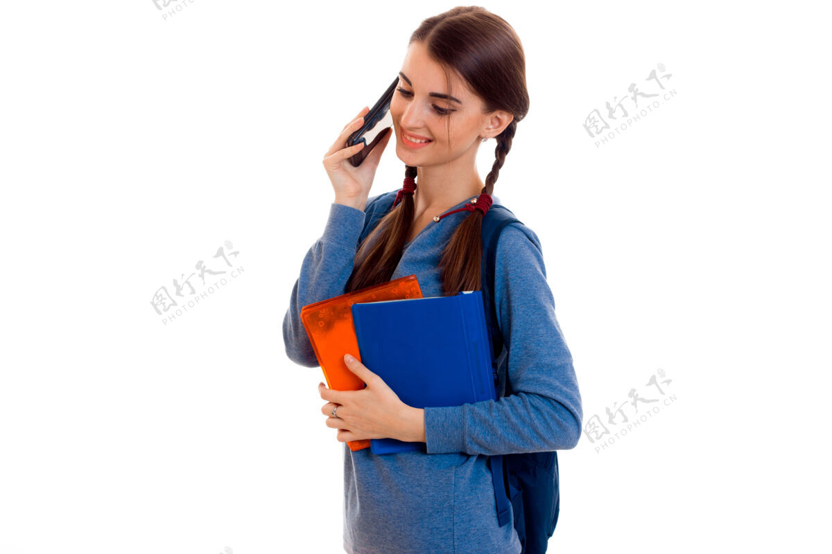 女人年轻漂亮的女学生背着背包在工作室的白墙上摆姿势和讲电话肖像乐趣快乐