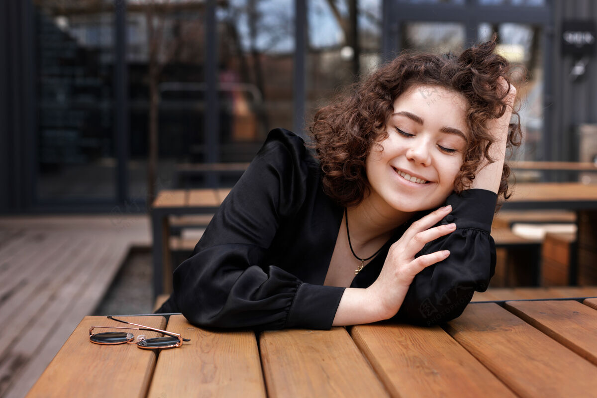 人穿着黑色夹克的快乐卷发女孩微笑着坐在街上的一张木桌旁坐着女人姿势