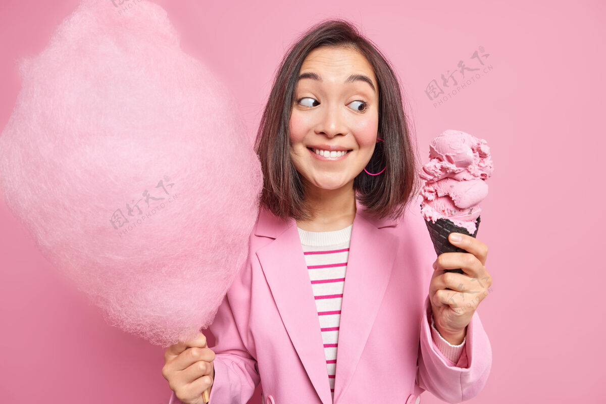 寒冷容貌讨人喜欢的黑发亚洲女人看着开胃的锥形冰淇淋拿着棉花糖放在棍子上享受夏日甜点吃垃圾食品穿着粉色夹克摆出室内有人在公园散步的姿势棉花漂亮姿势
