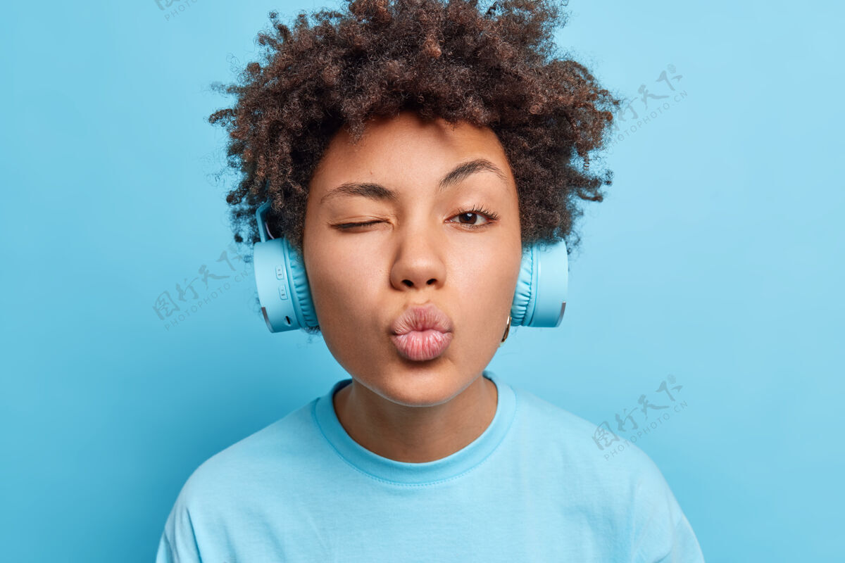 美国特写镜头深情可爱的非裔美国妇女眨眼眼睛保持嘴唇圆润喜欢通过耳机听音频轨道穿着随意隔离在蓝色的墙壁爱好的概念自然卷发满足