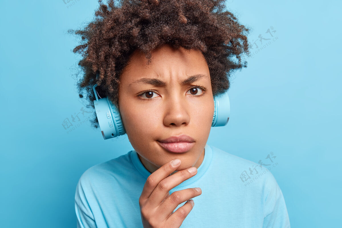 耳机严肃的非裔美国女孩的特写肖像手放在下巴上看起来不高兴穿着随便通过耳机听音乐或学习新的外国单词隔离在蓝色的墙壁享受人物人类
