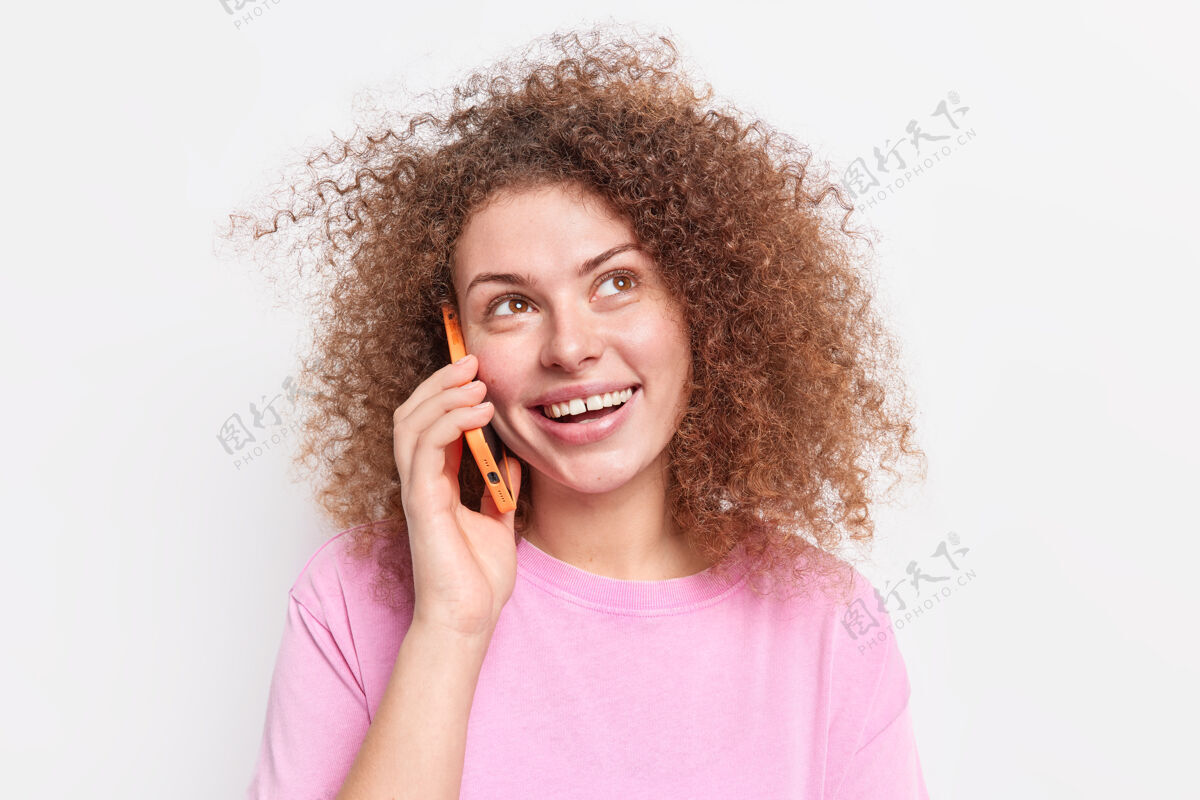 电话自然卷发的积极女人打电话给朋友见面拿着智能手机靠近耳朵高兴地向上看享受有趣的对话穿着休闲粉色t恤隔着白墙专注快乐设备