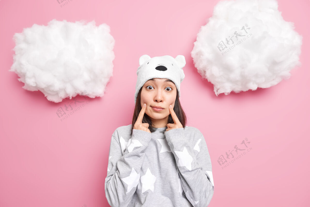手指女人微笑 手指靠近嘴角 戴着柔软的熊帽 休闲套头衫穿粉色看起来很孤立睡衣休闲穿