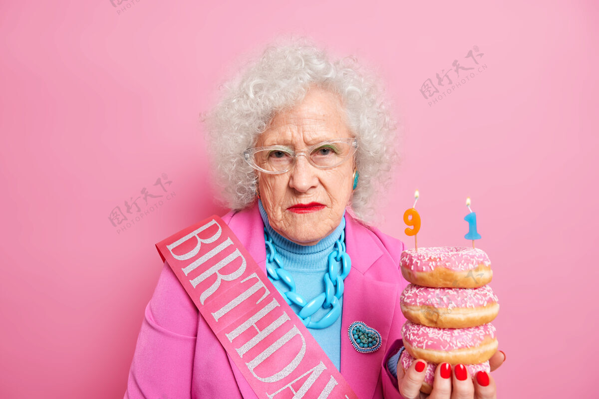 卷发摄影棚拍摄的满脸皱纹 头发卷曲的女人表情严肃 手里拿着一堆美味的油炸圈饼 穿着节日的衣服堆蛋糕粉色