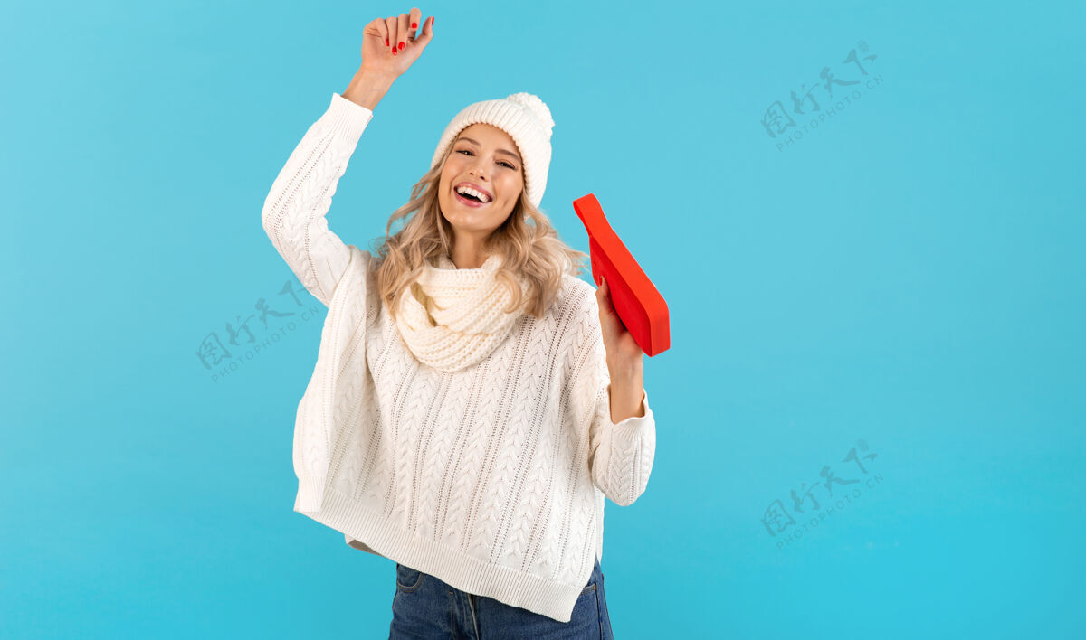 圣诞节时尚的金发微笑美丽的年轻女子手持无线扬声器听音乐快乐跳舞穿着白色毛衣和针织帽子冬季时尚摆造型毛衣配件衣服
