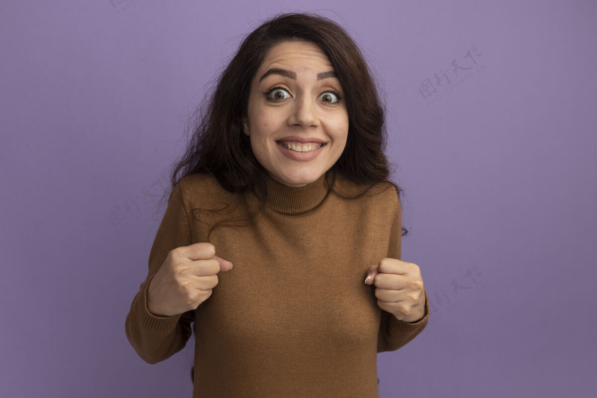 年轻兴奋的年轻漂亮女孩穿着棕色高领毛衣 在紫色的墙上显示“是”的手势站着高领毛衣是的