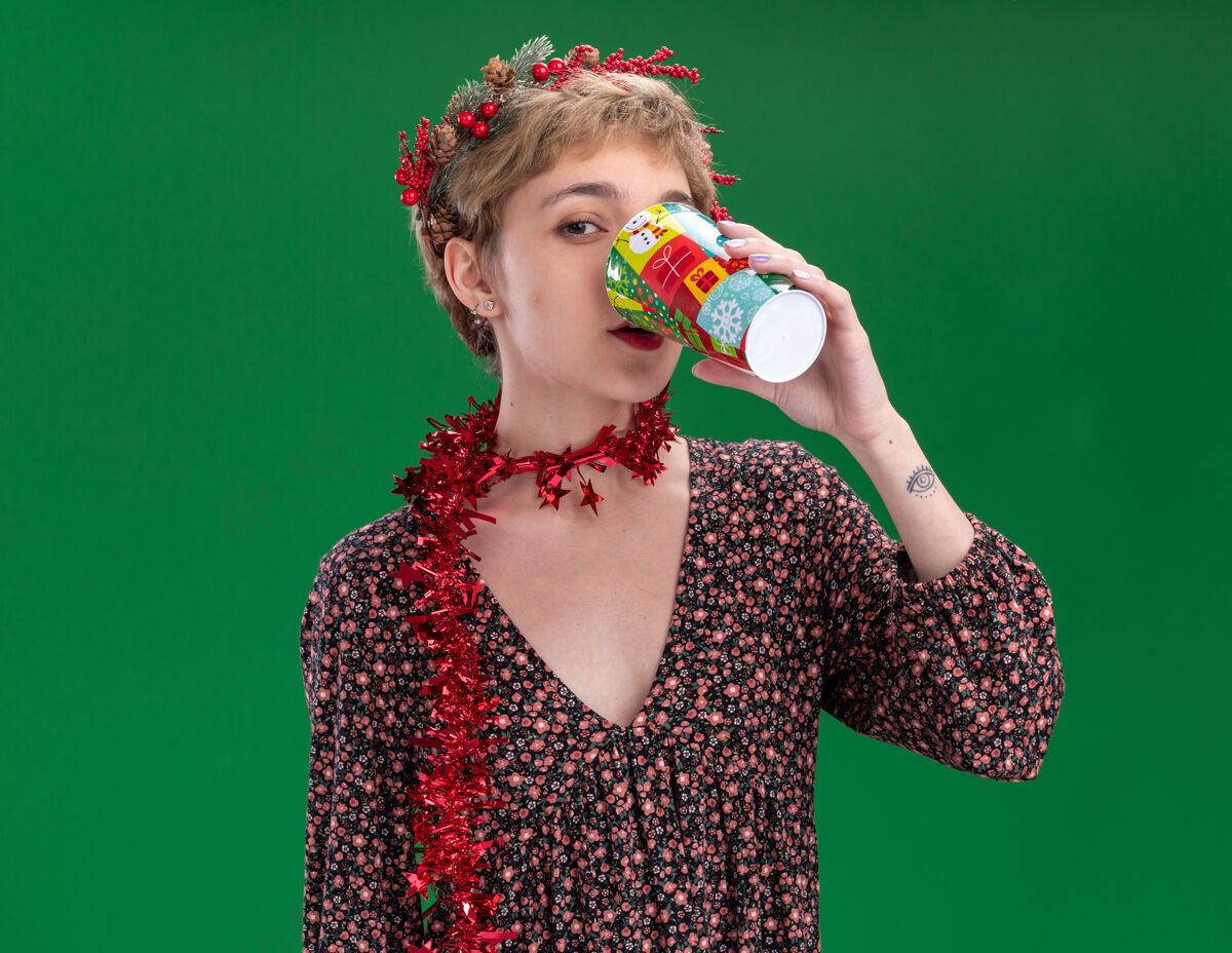 头年轻漂亮的女孩戴着圣诞花环 脖子上戴着金属丝花环 喝着塑料圣诞杯里的咖啡 这个杯子被隔离在绿色的墙上 有复制空间年轻咖啡脖子