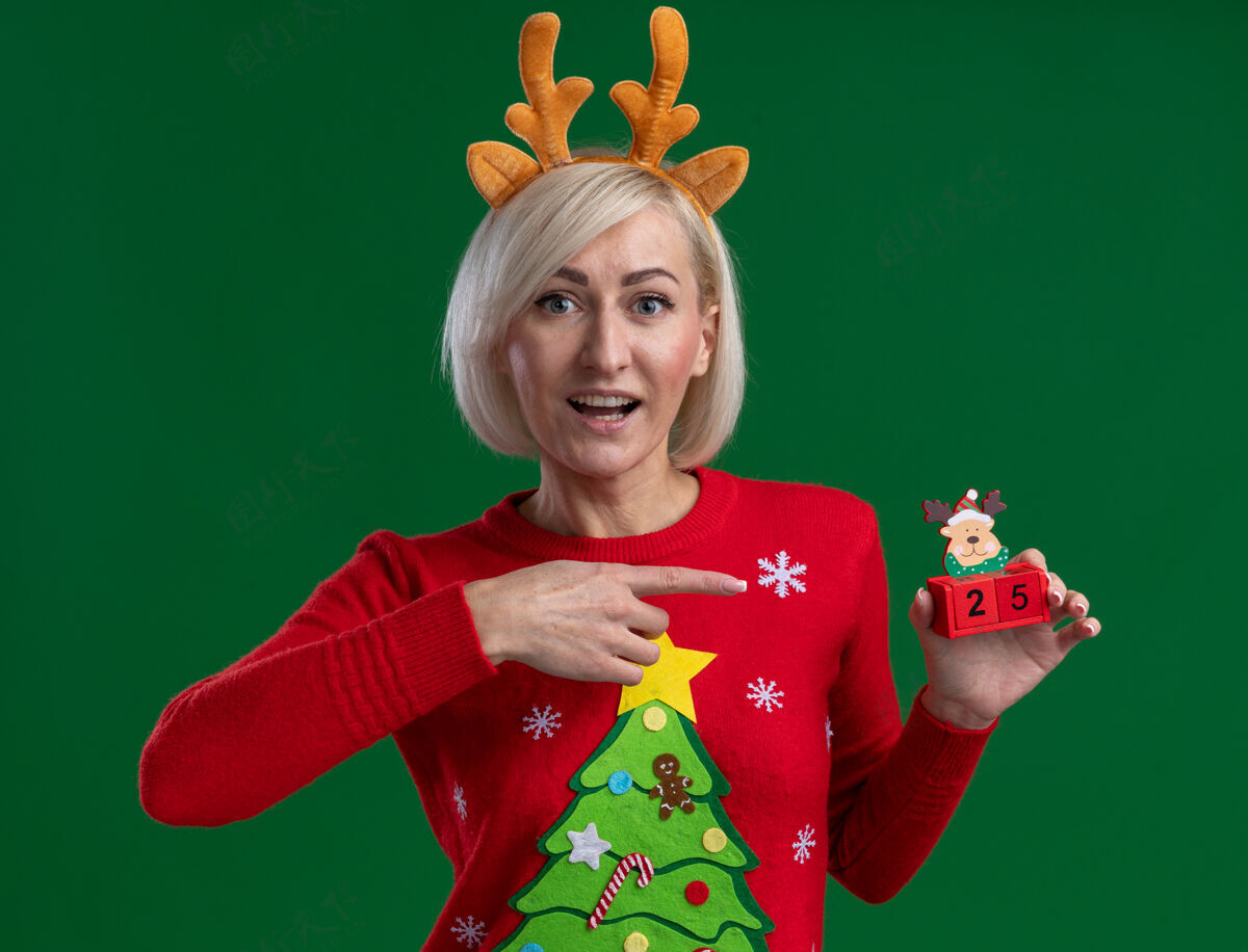 指着令人印象深刻的中年金发女人戴着圣诞驯鹿鹿角头带和圣诞毛衣拿着圣诞驯鹿玩具和日期看绿色墙上孤立印象驯鹿毛衣