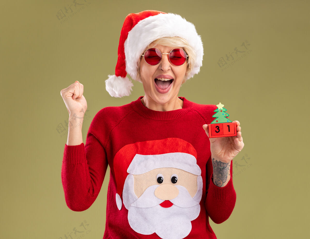 圣诞老人快乐的金发女郎戴着圣诞帽 穿着圣诞毛衣戴着圣诞眼镜拿着圣诞树玩具和日期做着“是”的手势 闭着眼睛孤立地站在橄榄绿的墙上穿圣诞节年轻