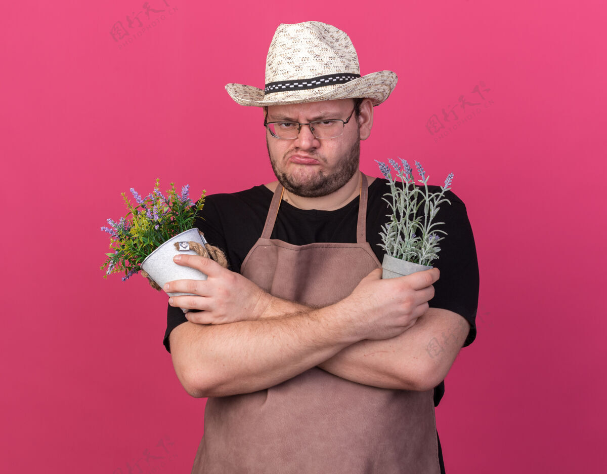 花园皱着眉头的年轻男园丁戴着园艺帽 拿着和粉色墙壁隔离的花盆里的花交叉脸举行皱眉