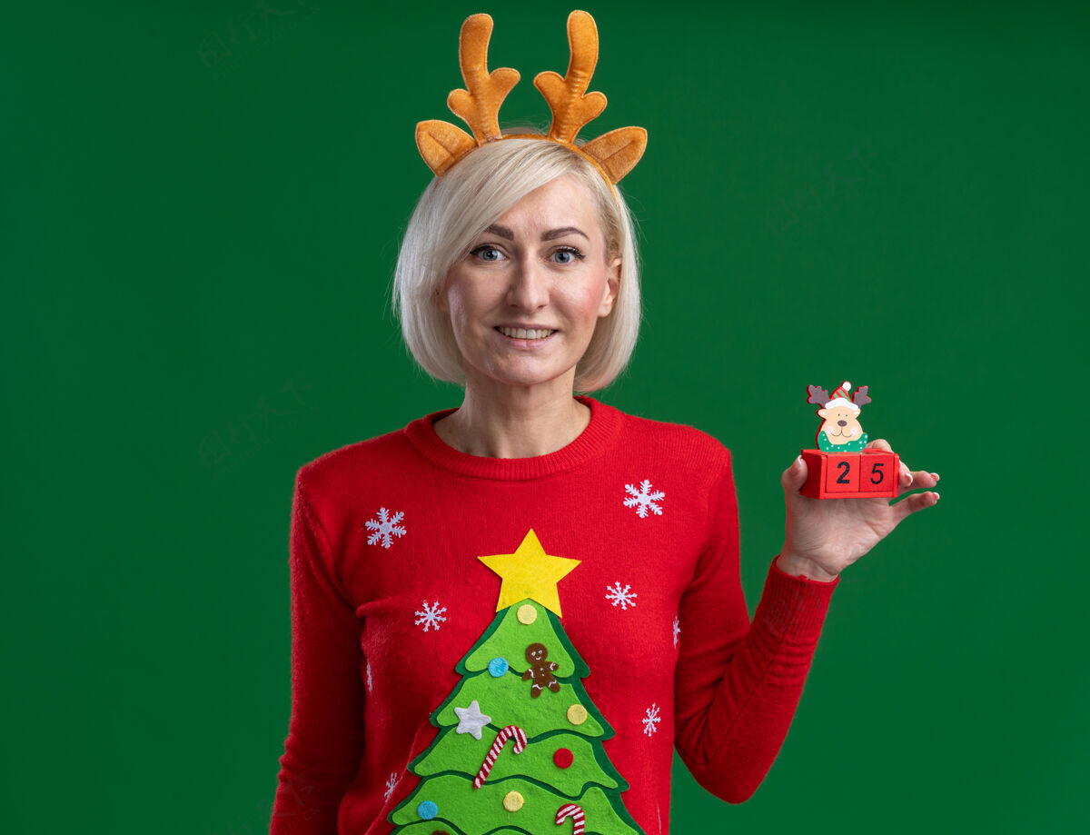 微笑微笑的中年金发女人戴着圣诞驯鹿鹿角头带和圣诞毛衣拿着圣诞驯鹿玩具和日期看起来孤立在绿色墙上的复制空间女人毛衣圣诞节