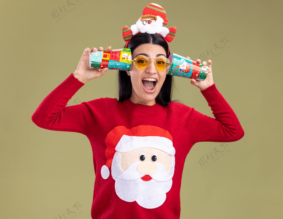圣诞老人紧张的白人女孩戴着圣诞老人的头带和毛衣戴着眼镜拿着塑料圣诞杯紧挨着耳朵听着谈话尖叫隔离在橄榄绿的墙上年轻杯子白种人