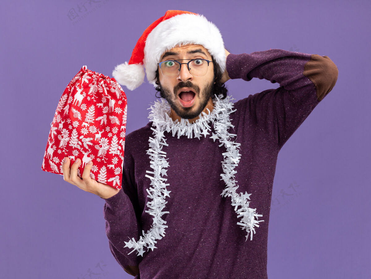 手年轻帅哥戴着圣诞帽 脖子上戴着花环 手里拿着圣诞包 手放在脑后 被隔离在蓝色的墙上帅气花环穿