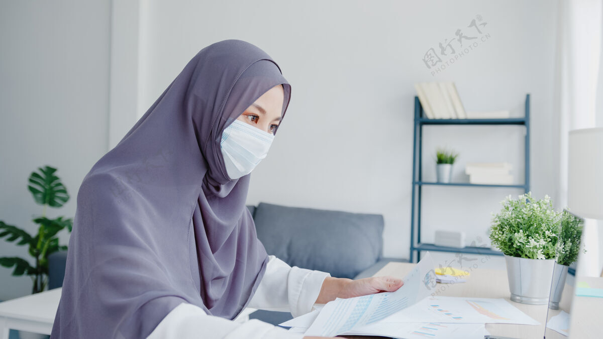 亚洲美丽的亚洲穆斯林女士戴着口罩在客厅使用笔记本电脑和商业报告穆斯林技术头巾