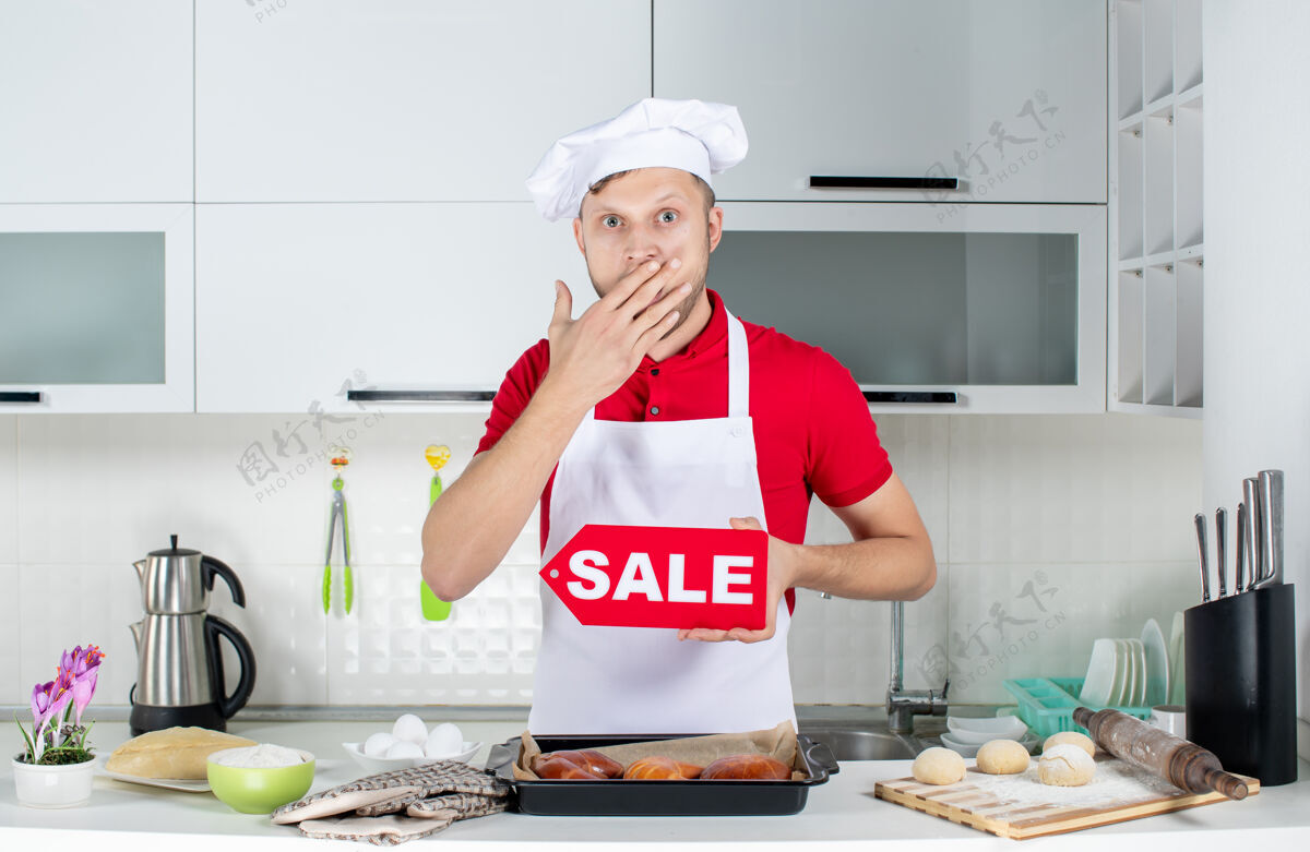 男人在白色厨房里 年轻惊讶的男厨师展示销售标志的俯视图工人年轻成人