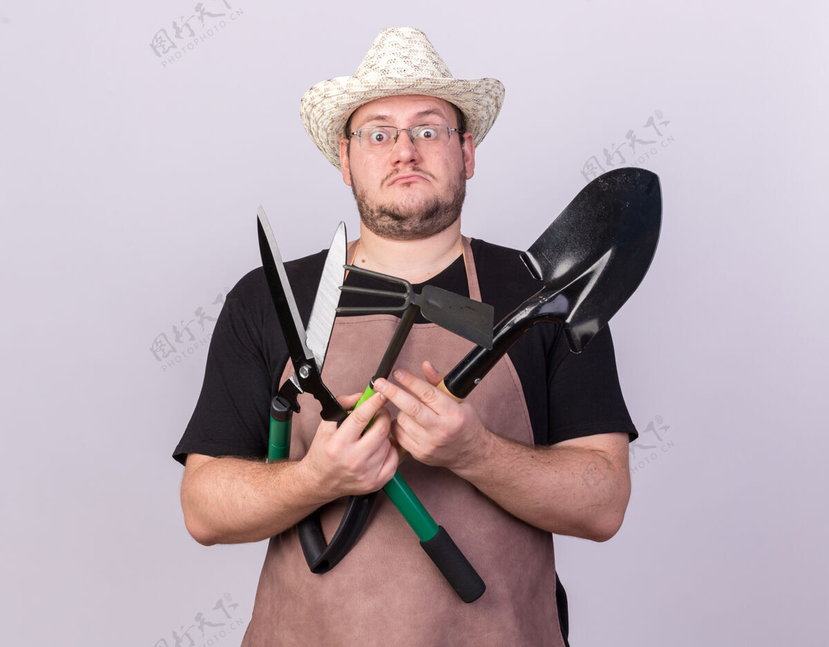 持有惊讶的年轻男园丁戴着园艺帽拿着铁锹用剪刀和锄头耙隔离在白色的墙上人男性锄头