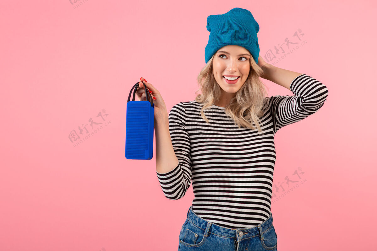 情感年轻漂亮的女人拿着无线扬声器听音乐穿着条纹衬衫和蓝色帽子微笑快乐积极的心情摆在粉红色的背景上积极衣服季节