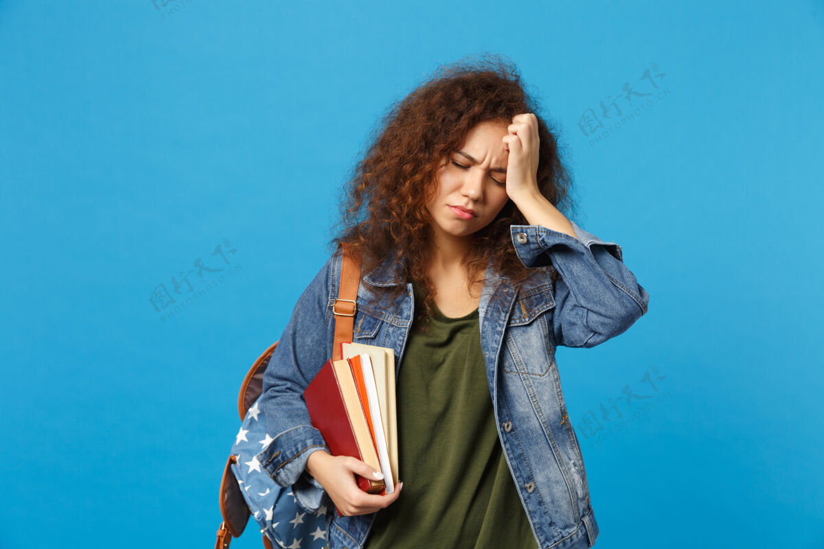 青少年穿着牛仔服 背着书包的悲伤的年轻学生把书孤立地放在蓝色的墙上人问题头痛